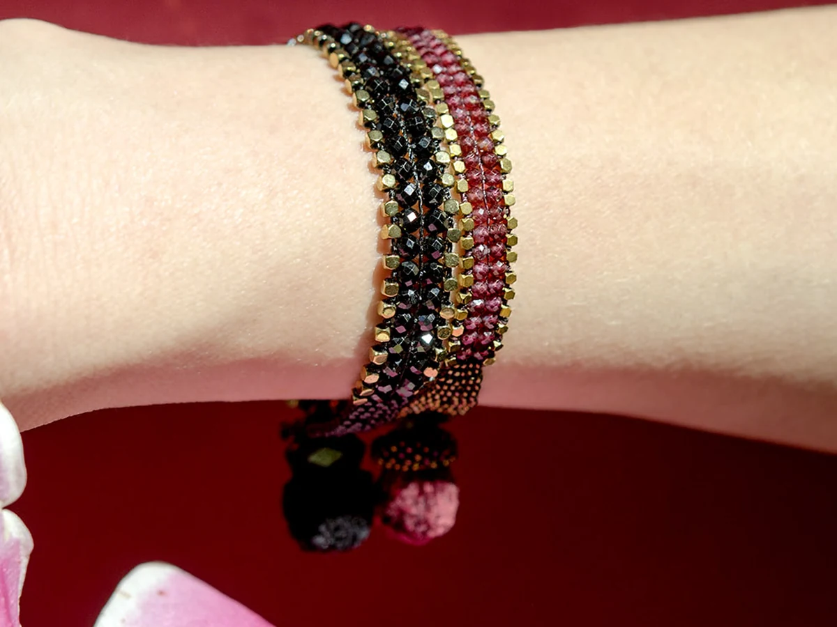 Black Spinel Gold Edge Bracelet and Garnet Black Gold Edge Bracelet stacked on a wrist, showing beaded detail and Oaxacan silk pompoms, against a deep red background.