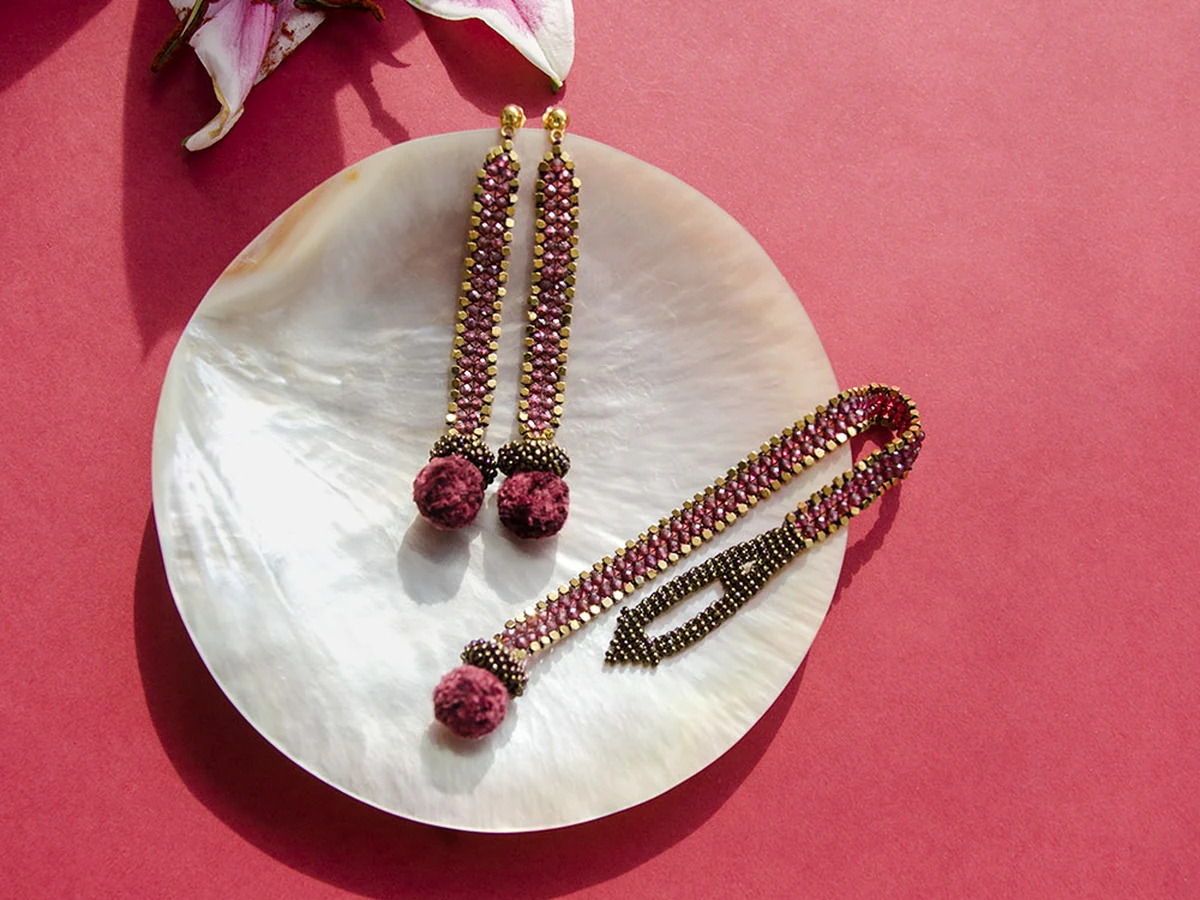 Atelier Lirio Garnet Gold Edge Earrings and a matching bracelet, made with garnet, Czech glass, hematite, displayed on a mother-of-pearl dish beside a pink lily, set against a red background.